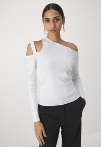 Top bianco a maniche lunghe con design a spalla singola, caratterizzato da piccole perforazioni su tutta la superficie e una texture a coste. Abbinato a pantaloni neri.