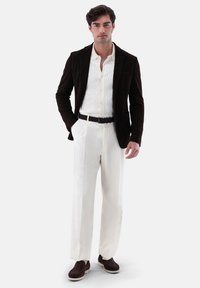 Blazer in velluto a coste marrone, camicia bianca con texture, pantaloni crema e scarpe marroni. Design chiaro con vestibilità sartoriale, dettagli con bottoni e accentuazione della cintura.