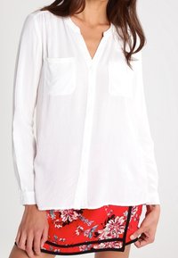 Blusa bianca a maniche lunghe con colletto a scollo a V, dettagli con tasche e chiusura con bottoni, abbinata a pantaloni corti rossi floreali con accenti neri.
