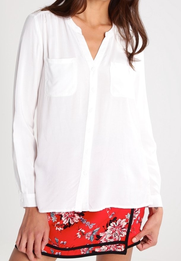 Blusa bianca a maniche lunghe con colletto a scollo a V, dettagli con tasche e chiusura con bottoni, abbinata a pantaloni corti rossi floreali con accenti neri.