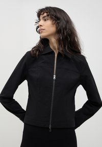 Donna con capelli lunghi e mossi di colore scuro, indossa una giacca nera con zip e colletto, in piedi con le mani dietro la schiena, mentre guarda di lato su uno sfondo neutro.