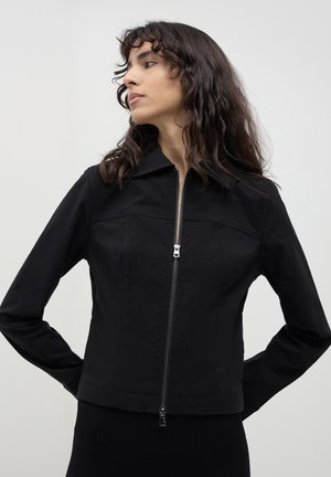 Eine Frau mit welligem, dunklem Haar trägt eine schwarze Zip-Up-Jacke mit Kragen, steht mit den Händen hinter ihrem Rücken und schaut zur Seite vor einem schlichten Hintergrund.