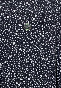 Námořnická látka s bílým abstraktním vzorem, doplněná stříbrným knoflíkem se zelenými akcenty a označením "Street One". Detail s plisováním.