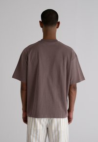 Brown cotton t-shirt with a relaxed fit, short sleeves, and a round neckline, paired with light striped pants. No notable details visible.