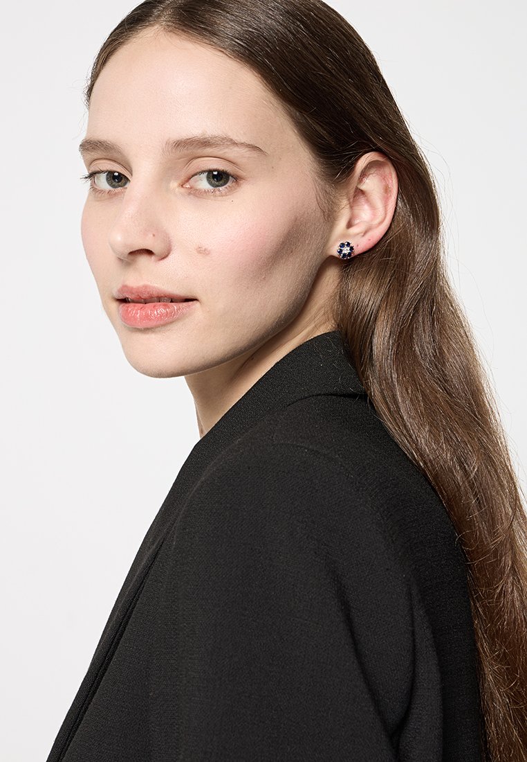 Ohrringe mit blauem Blumenmuster, runde Form, gefasst in silberfarbenem Metall. Das Modell hat lange braune Haare und trägt einen schwarzen Blazer.