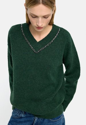 Kobieta ubrana w ciemnozielony sweter z dekoltem w kształcie litery V z ozdobami z kryształków, w połączeniu z niebieskimi jeansami, patrząca w dół na tle jednolitej ściany.