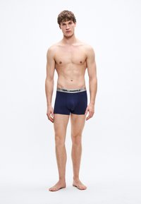 Bóxers azul marino hechos de tela suave, con una cinturilla plateada que lleva el texto "KARL LAGERFELD". El modelo está descalzo, con el torso descubierto.