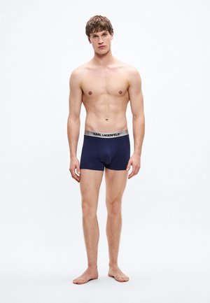 Bóxers azul marino hechos de tela suave, con una cinturilla plateada que lleva el texto "KARL LAGERFELD". El modelo está descalzo, con el torso descubierto.