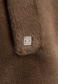 Tessuto in pelliccia marrone con una texture morbida presenta un'etichetta quadrata argentata, incisa con "Manila Grace". Metti in evidenza il colore naturale e i dettagli del materiale.