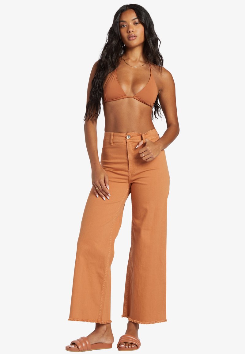 Donna con lunghi capelli scuri che indossa un top bikini color ruggine, pantaloni larghi a vita alta e sandali abbinate, in piedi con la mano sulla vita.