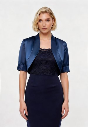 Γυναίκα που φοράει σατέν μπουφάν μπόλερο σε ναυτικό μπλε χρώμα πάνω από μαύρο δαντελένιο τοπ και στενή φούστα μολύβι σε ναυτικό μπλε χρώμα με ψηλή μέση, στέκεται μπροστά σε απλό φόντο.