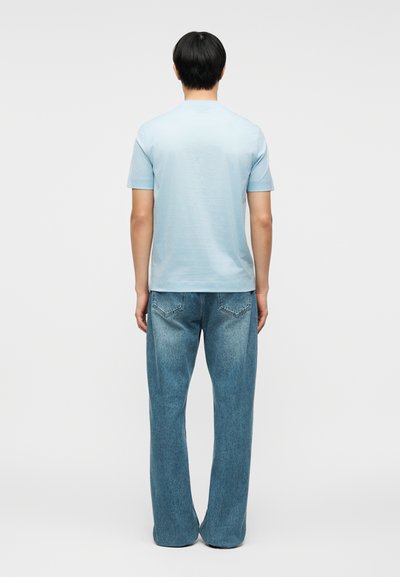 Άνδρας που στέκεται με την πλάτη γυρισμένη, φορώντας ανοιχτό μπλε κοντομάνικο πουκάμισο και άνετο μπλε τζιν, πάνω σε απλό φόντο.