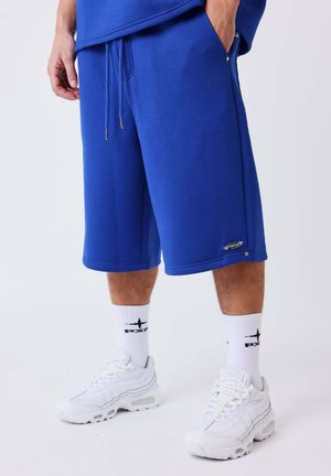 Man staat, draagt blauwe knielange shorts, witte sportsokken met zwart logo en witte sportschoenen tegen een effen lichte achtergrond.