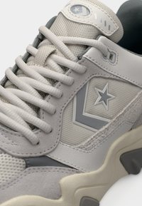 Gros plan d'une basket gris clair avec un tissu texturé, des panneaux en daim et cuir, un logo étoile et des lacets blancs épais sur une semelle épaisse.