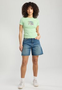 T-shirt ajustée vert clair avec un motif graphique, associée à un short en denim bleu. Le modèle porte des baskets blanches et des chaussettes blanches. Design simple.