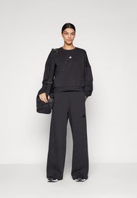 Juoda sportinė apranga, kurią sudaro trumpas džemperis ir plačios kelnės, abu su subtiliais logotipų akcentais. Papildyta maža juoda rankine.