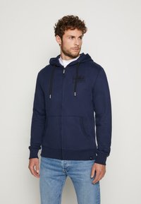 Marineblaue Zip-Up Kapuzenjacke aus weichem Baumwollmix, mit einer Fronttasche, verstellbarem Kordelzug an der Kapuze und kontrastierenden Nähdetails.