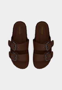 Καφέ σανδάλια με δύο ρυθμιζόμενους ιμάντες, που διαθέτουν μεταλλικά αγκράφες και υφή στη σόλα. Λείο υλικό με διακοσμητικά καρφιά.