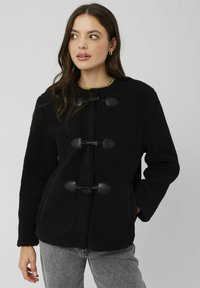 Veste en polaire noire avec un col rond, dotée de trois fermetures à nouer et de poches latérales. La texture est douce et moelleuse.