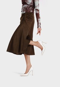 Gonna marrone, di lunghezza al ginocchio, con arricciature laterali, abbinata a scarpe con tacco alto e punta bianca. Il tessuto sembra liscio con una leggera lucentezza.