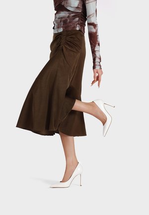 Gonna marrone, di lunghezza al ginocchio, con arricciature laterali, abbinata a scarpe con tacco alto e punta bianca. Il tessuto sembra liscio con una leggera lucentezza.