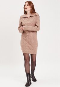 Robe en tricot beige clair avec un motif de tricot à torsades, un large col, une fermeture éclair à l'avant et des manches longues. Portée avec des bottines noires.