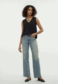 Donna con capelli ricci che indossa un top senza maniche nero, jeans a gamba larga blu chiaro, cintura nera, ballerine nere e una collana di grande impatto.