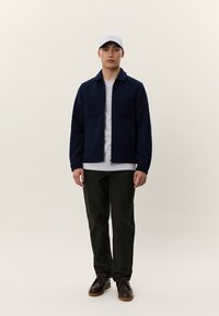 Ung mann iført grå caps, marineblå zip-up jakke, hvit skjorte, svarte bukser og mørkebrune sko, stående mot en ensfarget lys bakgrunn.