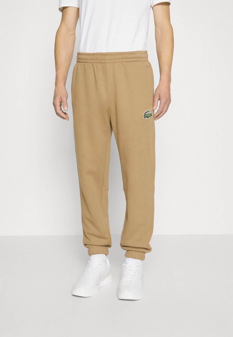 Braune Sweatpants mit elastischem Bund, Seitentaschen und einem grünen gestickten Logo am Oberschenkel, getragen mit weißen Turnschuhen.