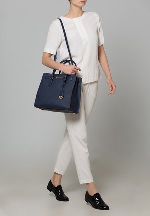 Femme portant une tenue blanche, tenant un grand sac à main bleu marine avec une bandoulière, et des mocassins noirs brillants, sur un fond gris uni.