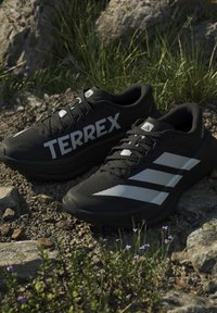 Двойка черни маратонки Adidas Terrex за бягане по пресечен терен с бяло лого на чакъл и скали близо до трева и малки лилави цветя.
