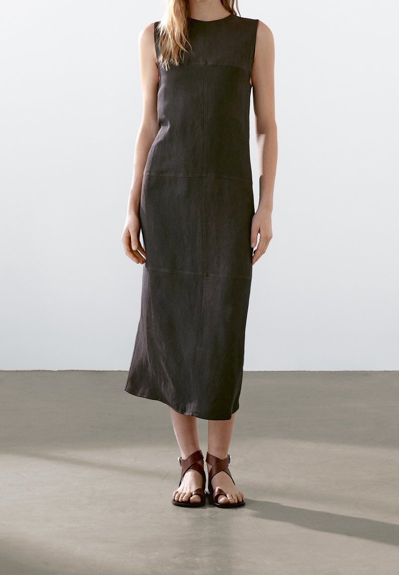 Femme portant une robe midi sombre sans manches et des sandales plates ouvertes marron, debout sur un sol lisse devant un mur clair.