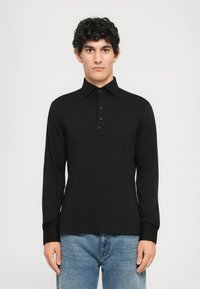 Ung mann med krøllete mørkt hår iført en svart langermet polo skjorte og blåjeans, stående mot en enkel hvit bakgrunn.