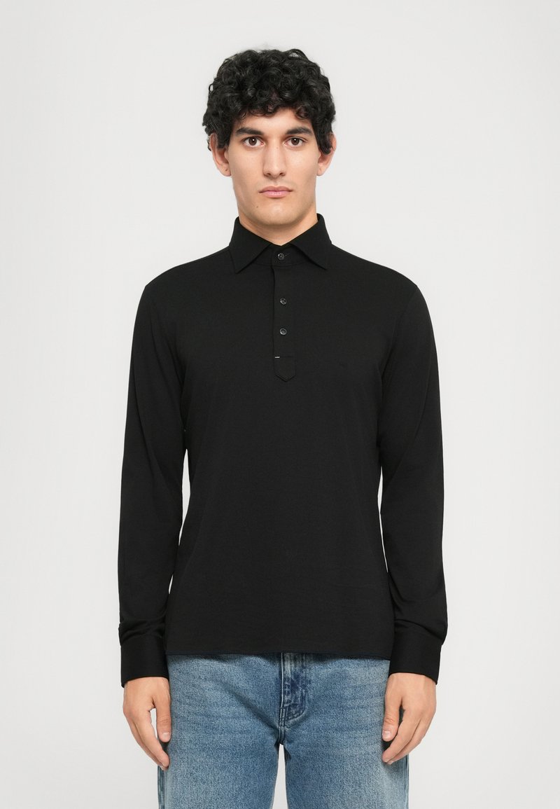 Ung mann med krøllete mørkt hår iført en svart langermet polo skjorte og blåjeans, stående mot en enkel hvit bakgrunn.