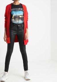 Person wearing a red cardigan, black graphic T-shirt, black leather pants, and white sneakers, standing against a plain white background.