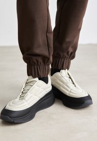 Beige vadderade sneakers med en svart gummiklädd sula och accent. Bärs med bruna uppvikta joggers som har en slät yta och en elegant design.