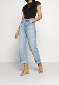 Czarny body z przezroczystymi, falbanianymi rękawami, zestawiony z jasnoniebieskimi jeansami z wysokim stanem i beżowymi sandałami na słupku.