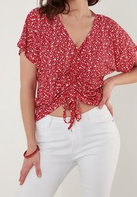 Blouse florale rouge à manches courtes, avec un décolleté en V et un détail froncé à la taille, associée à un jean blanc. Texture de tissu lisse.