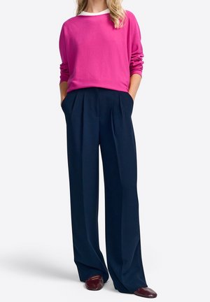 Vrouw in een felroze lange mouw top, marineblauwe wijde broek met plooien, handen in de zakken, en donkerrode platte schoenen.
