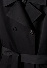 Détail d'un trench-coat noir montrant des boutons à double boutonnage, un large col à revers et une ceinture en tissu nouée à la taille.