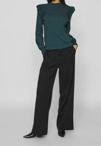 Femme portant un pull côtelé vert foncé avec des volants aux épaules et un pantalon noir ample, accompagné de chaussures noires à bouts pointus.