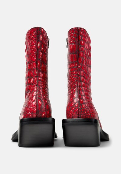 Bottines en cuir rouge à motif crocodile avec talons carrés noirs, montrées de dos sur un fond blanc.