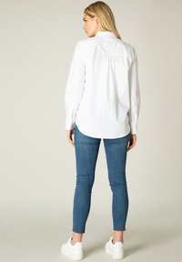 Witte lange-sleeve shirt met een afgeronde zoom en achterin, gecombineerd met een strakke blauwe jeans en witte sneakers, van achteren weergegeven.