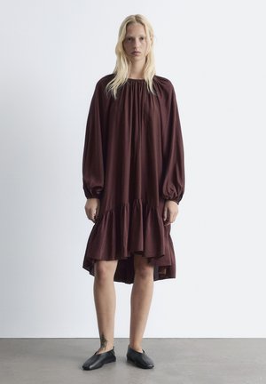 Burgunderrotes Langarmkleid mit gerafftem Ausschnitt und gestuftem Saum, aus fließendem Stoff in lockerem Schnitt. Tragen mit schwarzen flachen Schuhen.