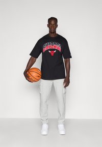 T-shirt oversize nera con "Chicago" e un logo di un toro, abbinata a pantaloni felpati grigi chiari e sneakers bianche, mentre tiene un pallone da basket.