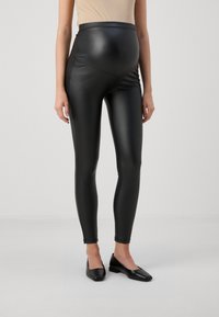 Graviditetstights i svart konstläder, högmidjad design, slät textur, åtsittande silhuett, i kombination med svarta ballerinaliknande skor, minimalistisk stil.