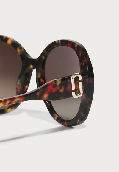 Schildpatt-Sonnenbrille mit dunklen Gläsern; massives Gestell mit goldenen Hardware-Details und glänzendem Finish. Akzente in Bernstein und Rot.