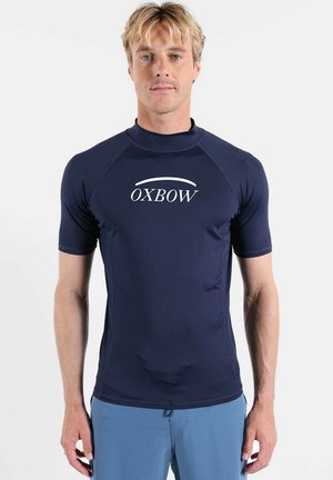 Homme blond portant un rash guard bleu marine à manches courtes avec le logo "OXBOW" et un short de bain bleu clair, debout devant un fond blanc.