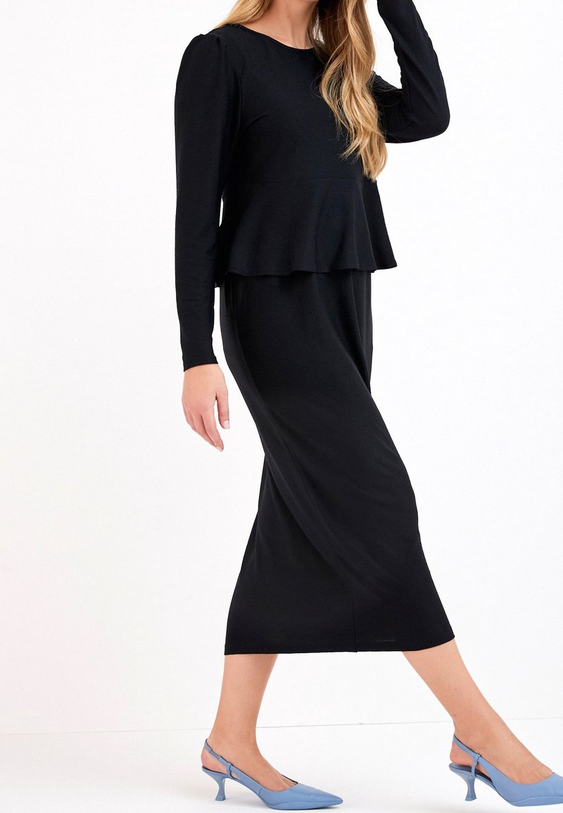Femme portant une longue robe noire à manches longues avec un détail peplum, associée à des escarpins slingback bleus pointus.