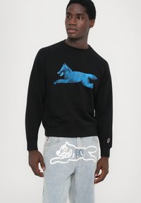 Black sweatshirt with blue graphic of a leaping wolf, paired with light blue jeans featuring a white wolf outline logo on the back pocket.